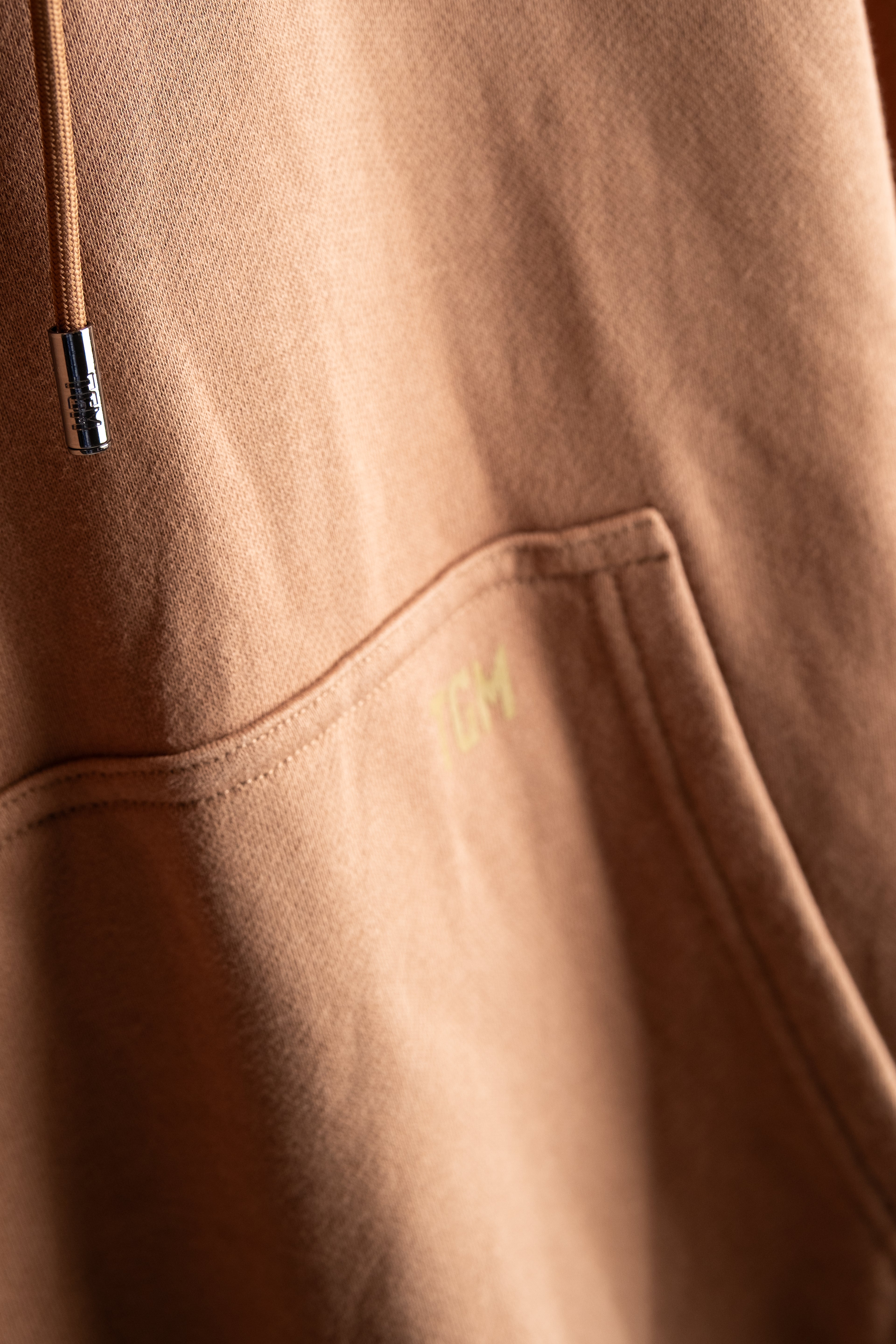 Close-up of a brown hoodie with a TGM brand logo on the sleeve. 100% premium cotton