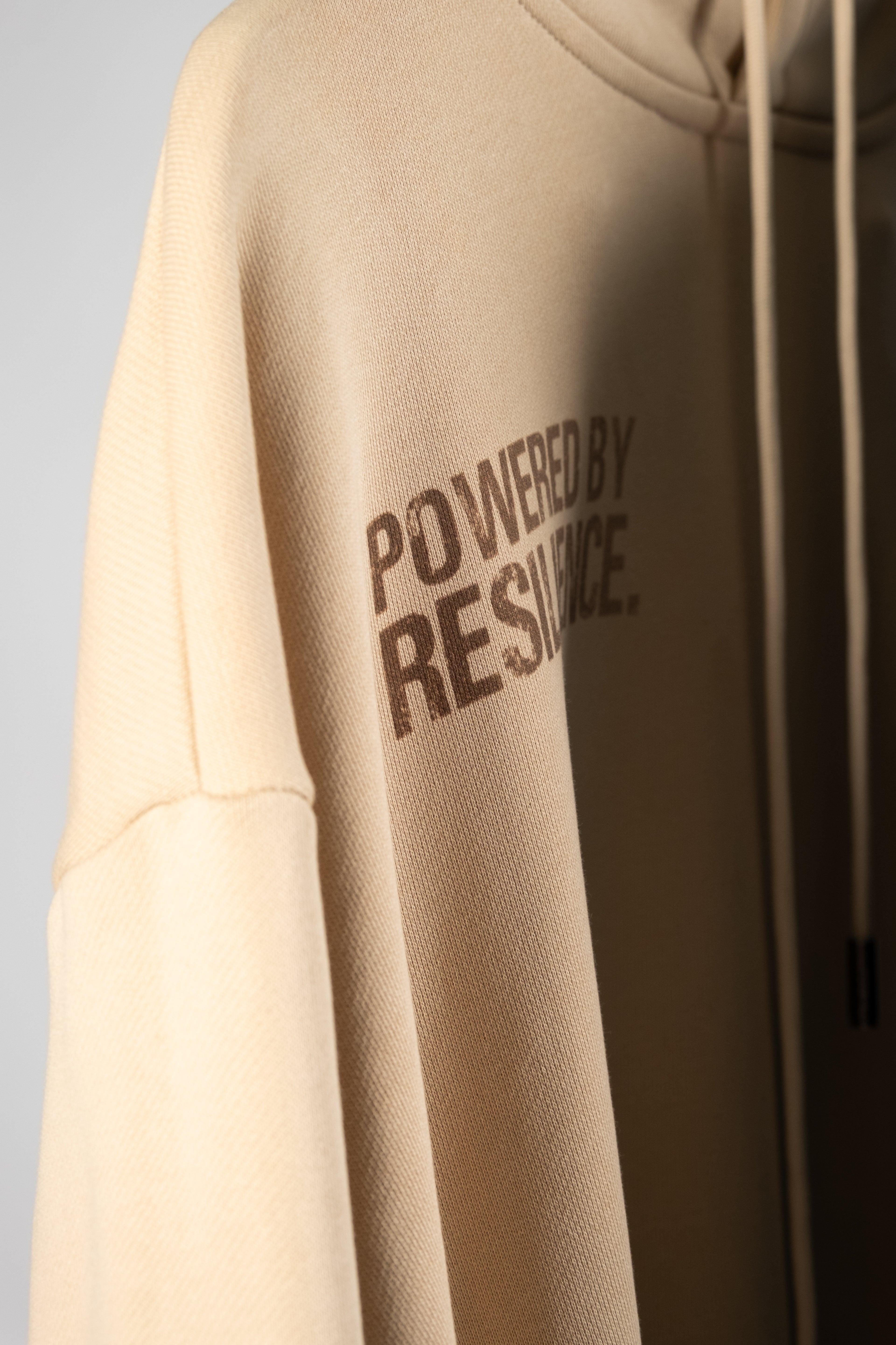 Beige Oversized Hoodie with brown gradient text saying powered by resilience close up of text. 100% premium cotton