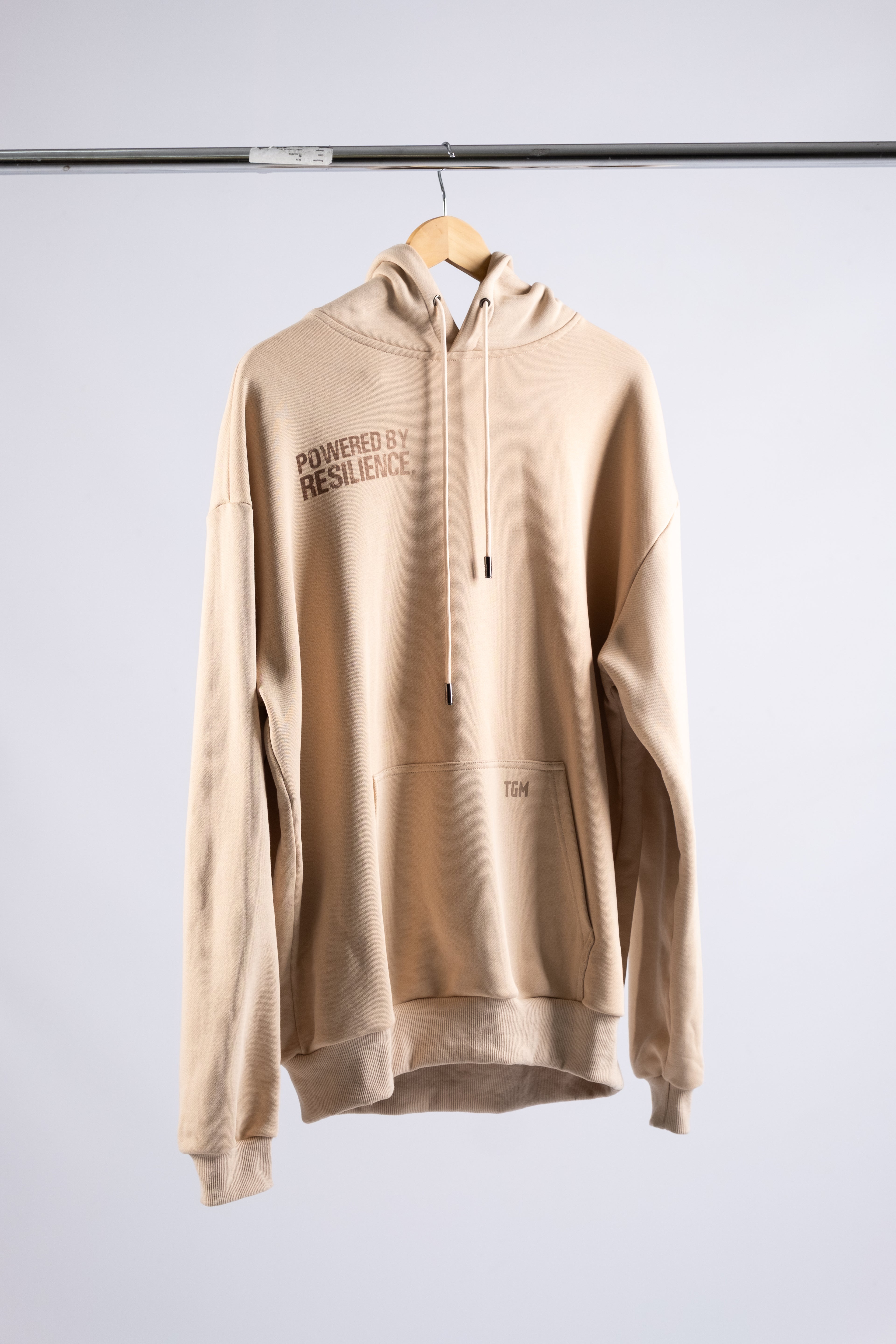 Beige Oversized Hoodie with brown gradient text saying powered by resilience. Hung on a hanger 100% premium cotton