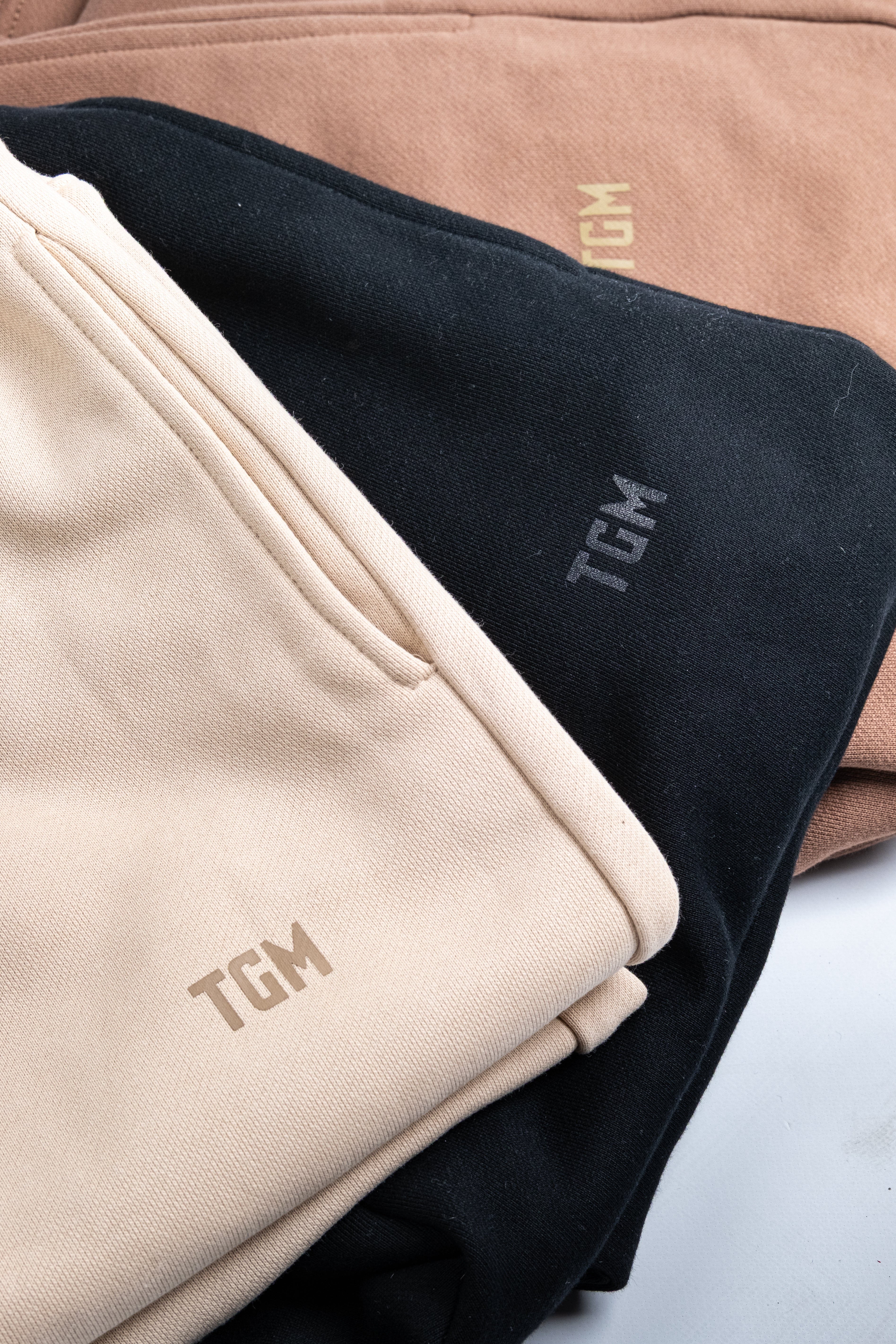 Folded sweatpants pieces in black, beige, and brown with 'TGM' branding.